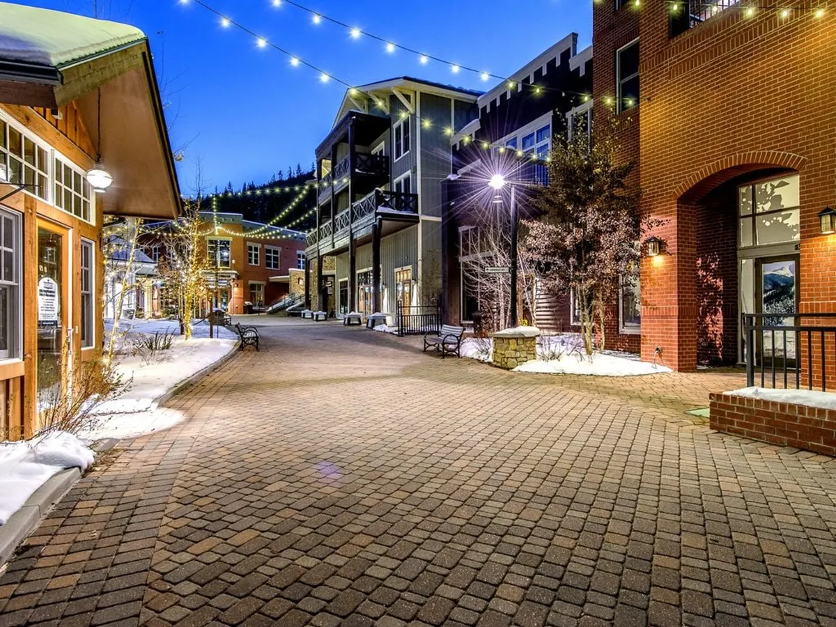 A lively base village scene illuminated by string lights, with a cobblestone pathway, benches, and festive seasonal décor. People are seen strolling, socializing, and enjoying the communal atmosphere, suggesting year-round vibrancy beyond winter sports.