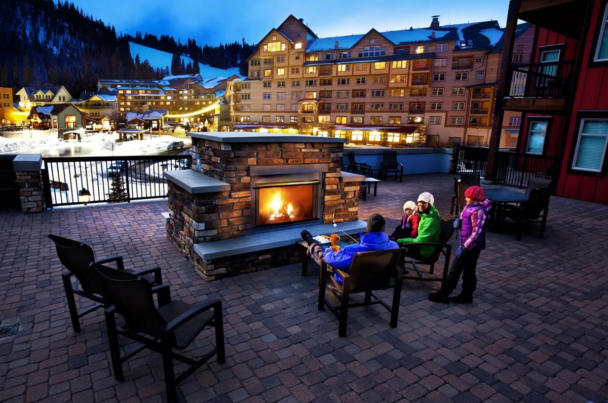 An inviting exterior scene featuring an outdoor fireplace surrounded by comfortable seating, with a beautiful mountain backdrop and an illuminated building facade in the evening. Families and friends can be seen gathering around the fire, enjoying warm drinks and sharing stories.