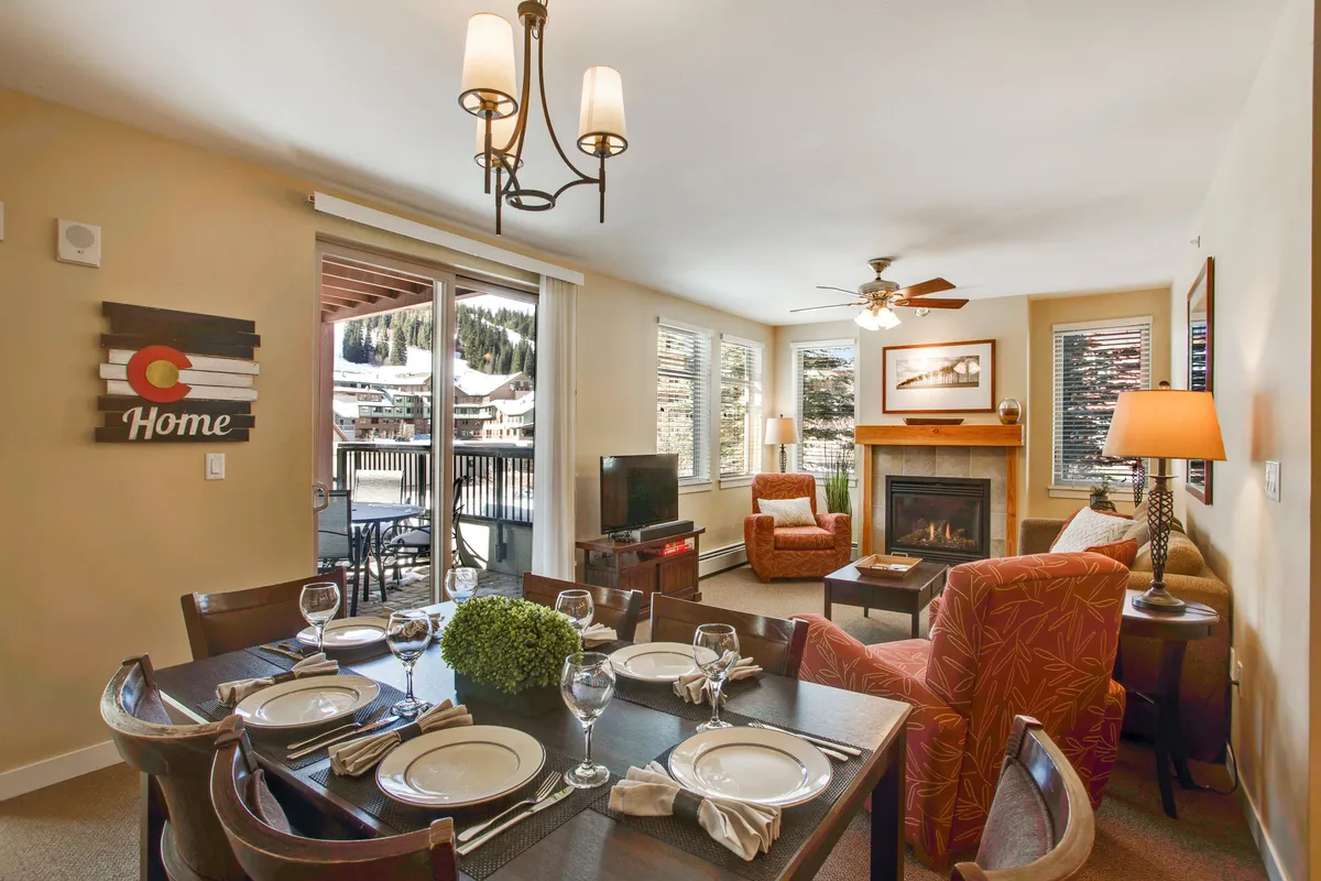This image features a warm, open-concept living area with a dining table, comfortable seating, a glowing stone fireplace, and expansive windows revealing scenic mountain views. The space is designed for group gatherings, with cozy furnishings and natural light enhancing the inviting atmosphere.
