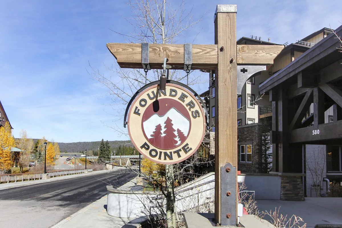 An exterior shot featuring the Founder's Pointe sign, framed by leafy trees and nearby buildings. The relaxed, inviting mood hints at the welcoming atmosphere guests will experience on arrival. The backdrop includes mountain vistas and a sense of connection to the broader property environment.