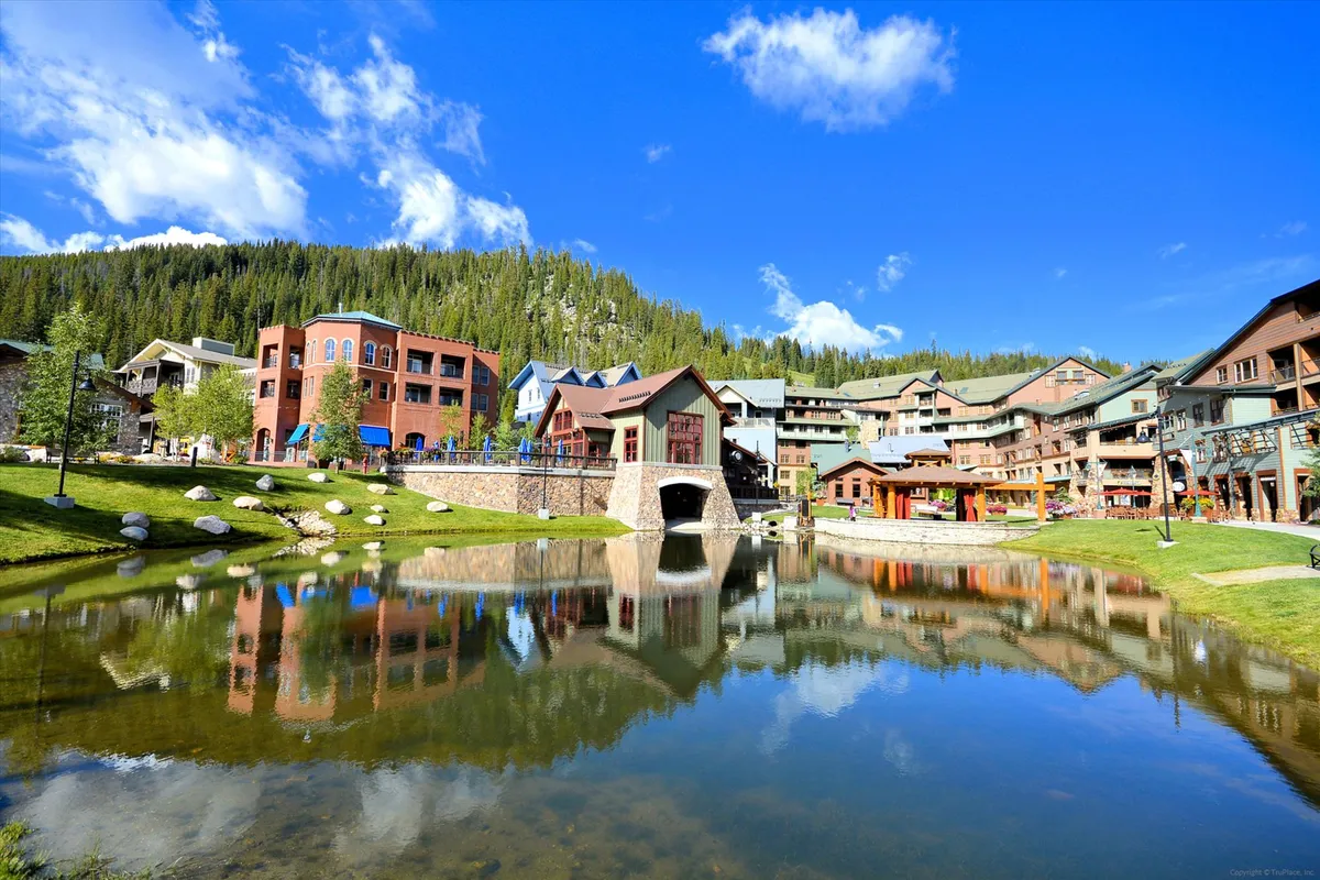 A colorful, cheerful village scene with buildings reflected in a serene pond, a pedestrian bridge, and lush trees. People are strolling, shopping, and enjoying the outdoor atmosphere, evoking a sense of vibrant community life.