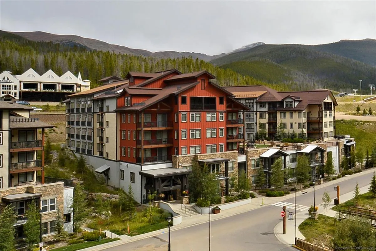 A welcoming exterior shot of a multi-story building nestled among trees with a majestic mountain backdrop. Guests are seen relaxing outdoors, exploring nearby trails, and socializing in communal areas, conveying a sense of easy access to adventure and community.