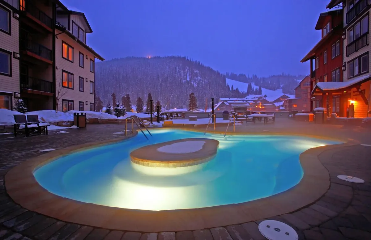 This image displays a heated outdoor pool and hot tub set against a snow-covered landscape, with illuminated building windows adding a cozy ambiance. People are seen relaxing in the hot tub, socializing, and enjoying the contrast of warm water with the winter air under the night sky.