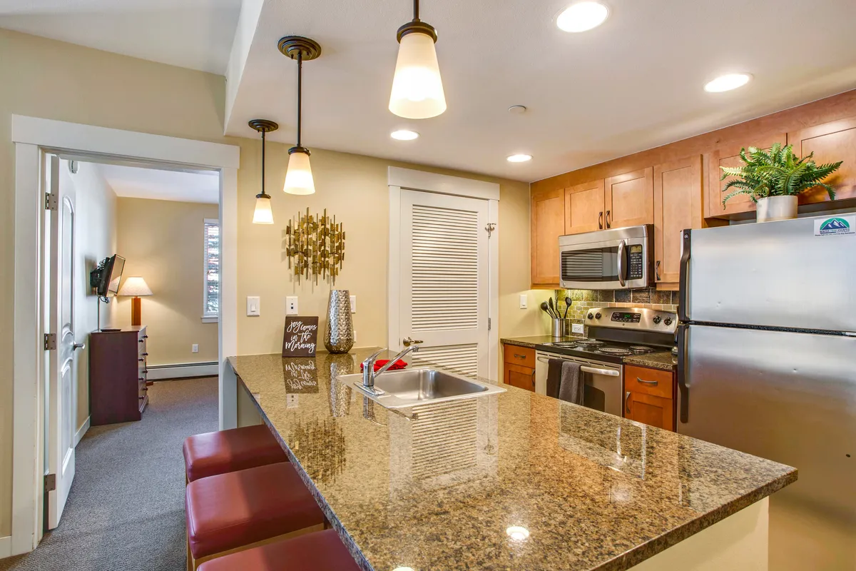 This image features a granite countertop, bar stools, modern kitchen appliances, and decorative wall art. The arrangement suggests a social atmosphere, where cooking, entertaining, and casual dining blend seamlessly. The lighting and décor add warmth, making the space feel both practical and inviting.