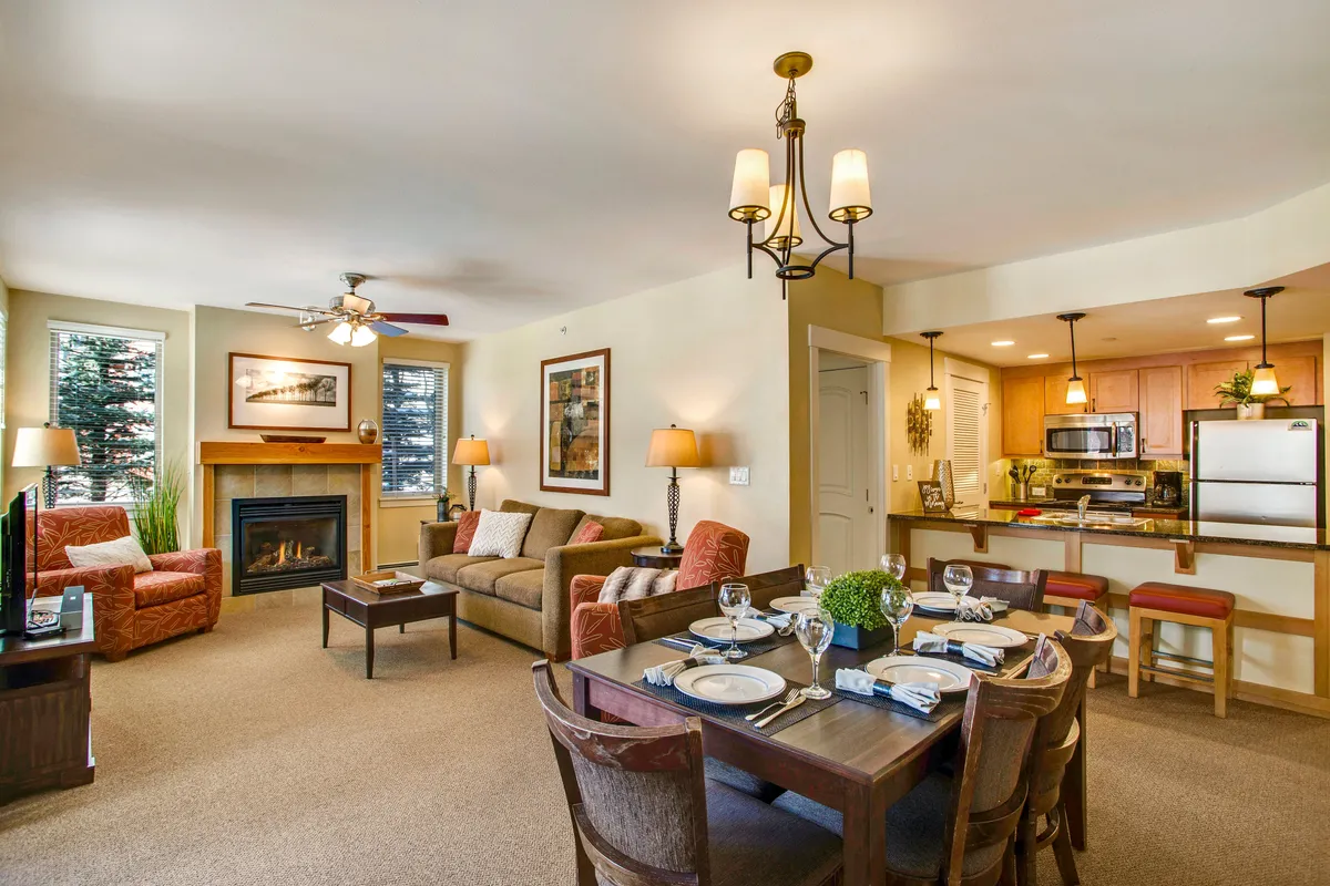 The image captures a warm, inviting living space with a glowing fireplace, a spacious dining table ready for shared meals, a plush sofa for relaxation, and a modern kitchen island. The interior exudes a cozy atmosphere perfect for gathering after a day on the slopes.