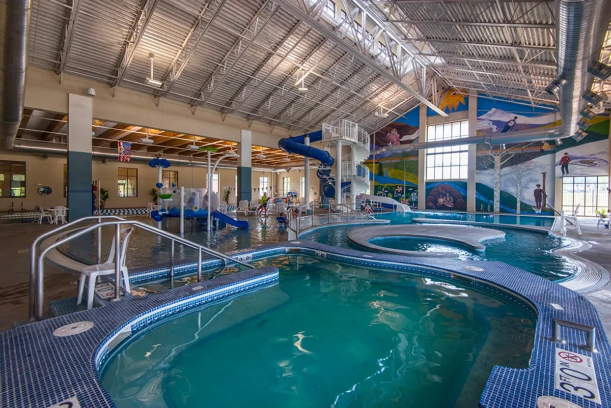 A lively indoor recreation area with a water slide, a meandering lazy river, poolside lounge chairs, and vibrant murals. Children and families are having fun swimming, sliding, and relaxing by the pool.