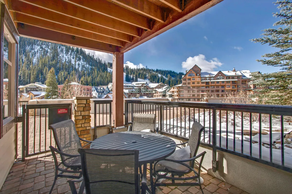 An inviting outdoor patio with a table and chairs surrounded by trees, neighboring buildings, and a breathtaking mountain view. The setup hints at outdoor dining, relaxing, or gathering with friends while soaking in serene alpine scenery.